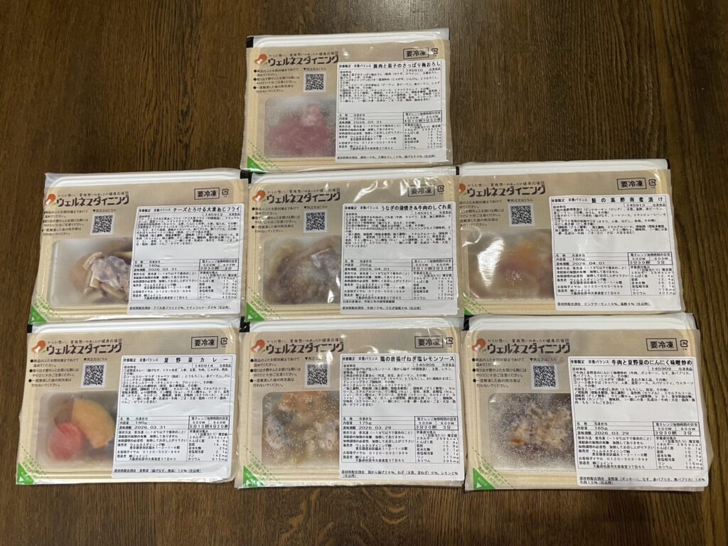 宅配された7種類の冷凍弁当がテーブルに並べられている様子。それぞれ主菜と副菜が分かれてパッケージされ、メニューのバリエーションが一目でわかる構成。